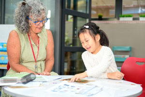 Young smiling student points at schoolwork while sitting at table with teacher.