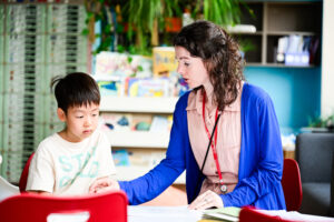 Teacher sitting and reading with young student at a table in the library. 