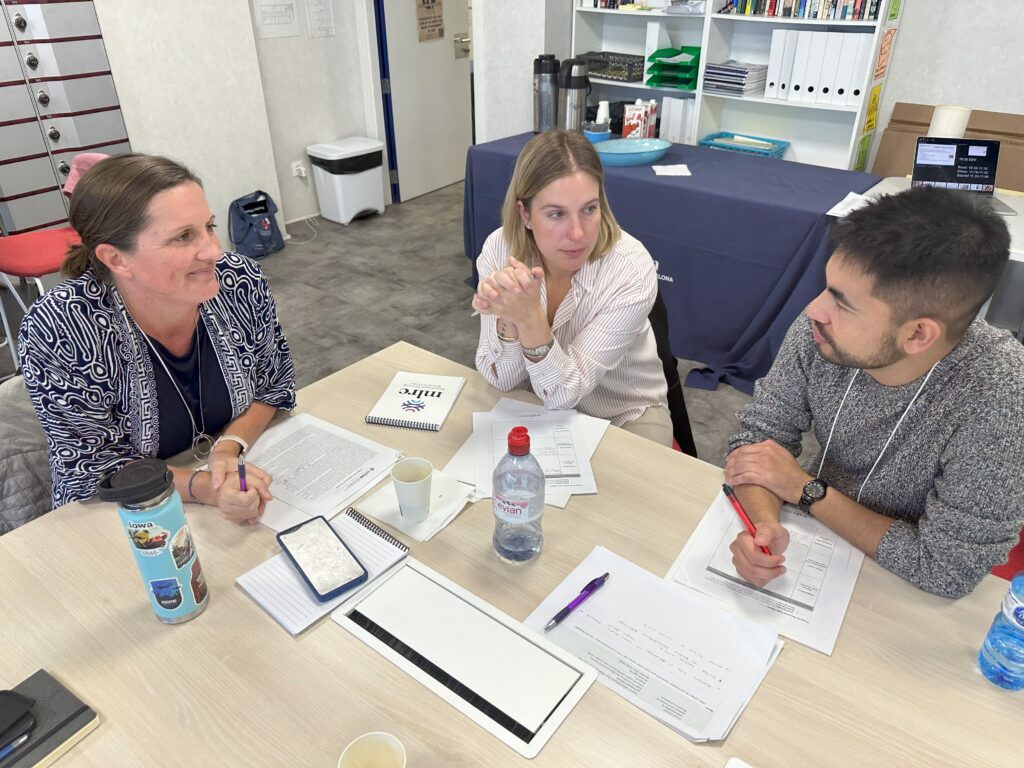 Three people having a discussion at a table that has notebooks and handouts on it.